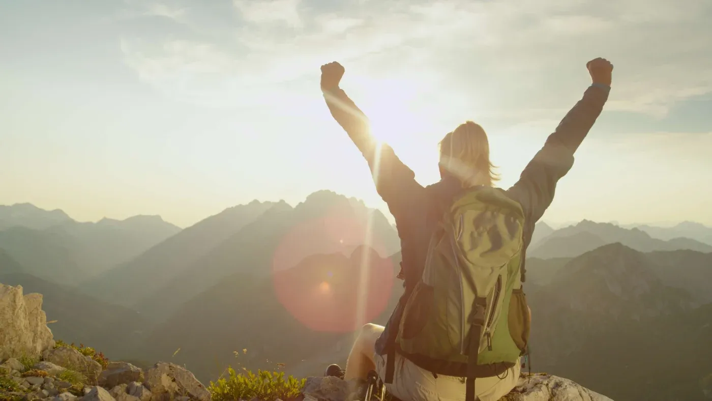 A hiker with a backpack sits on a rocky peak, arms raised triumphantly against a glowing sunrise, overlooking misty mountains. A sense of achievement fills the scene.