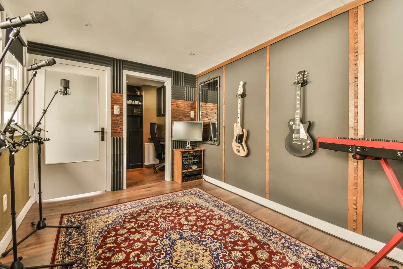 Home music studio with soundproofed walls features guitars hanging, microphones, a computer setup, a red keyboard, and a patterned rug on wooden floors.