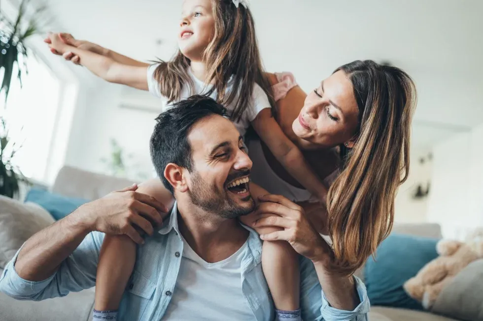 A joyful family moment in a bright living room. A father laughs while carrying a young girl on his shoulders. The mother smiles warmly at both.