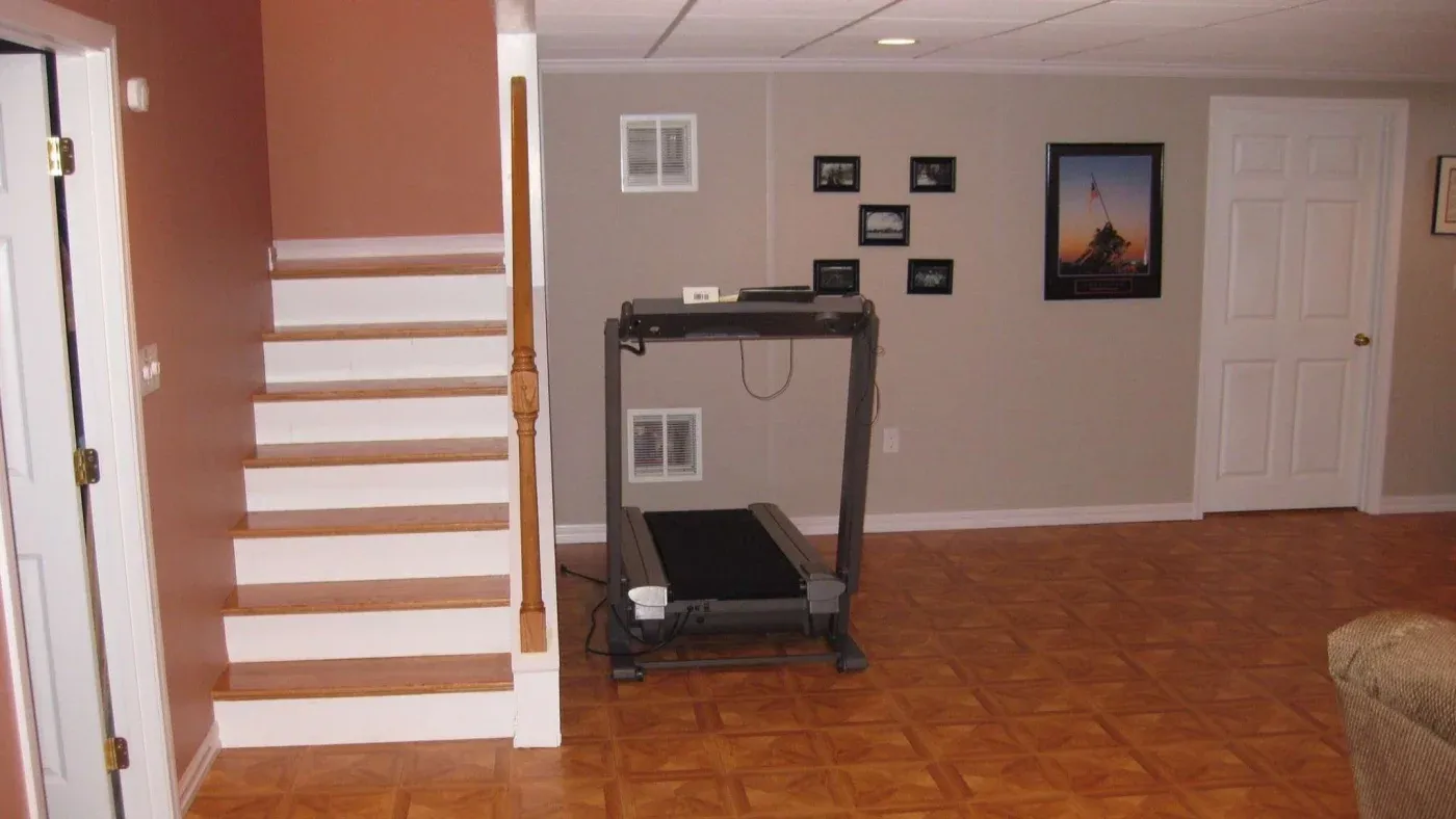A basement room with a treadmill against a beige wall adorned with small framed photos. To the left, a staircase with wooden steps ascends, and the floor is parquet. The atmosphere is tidy and functional.