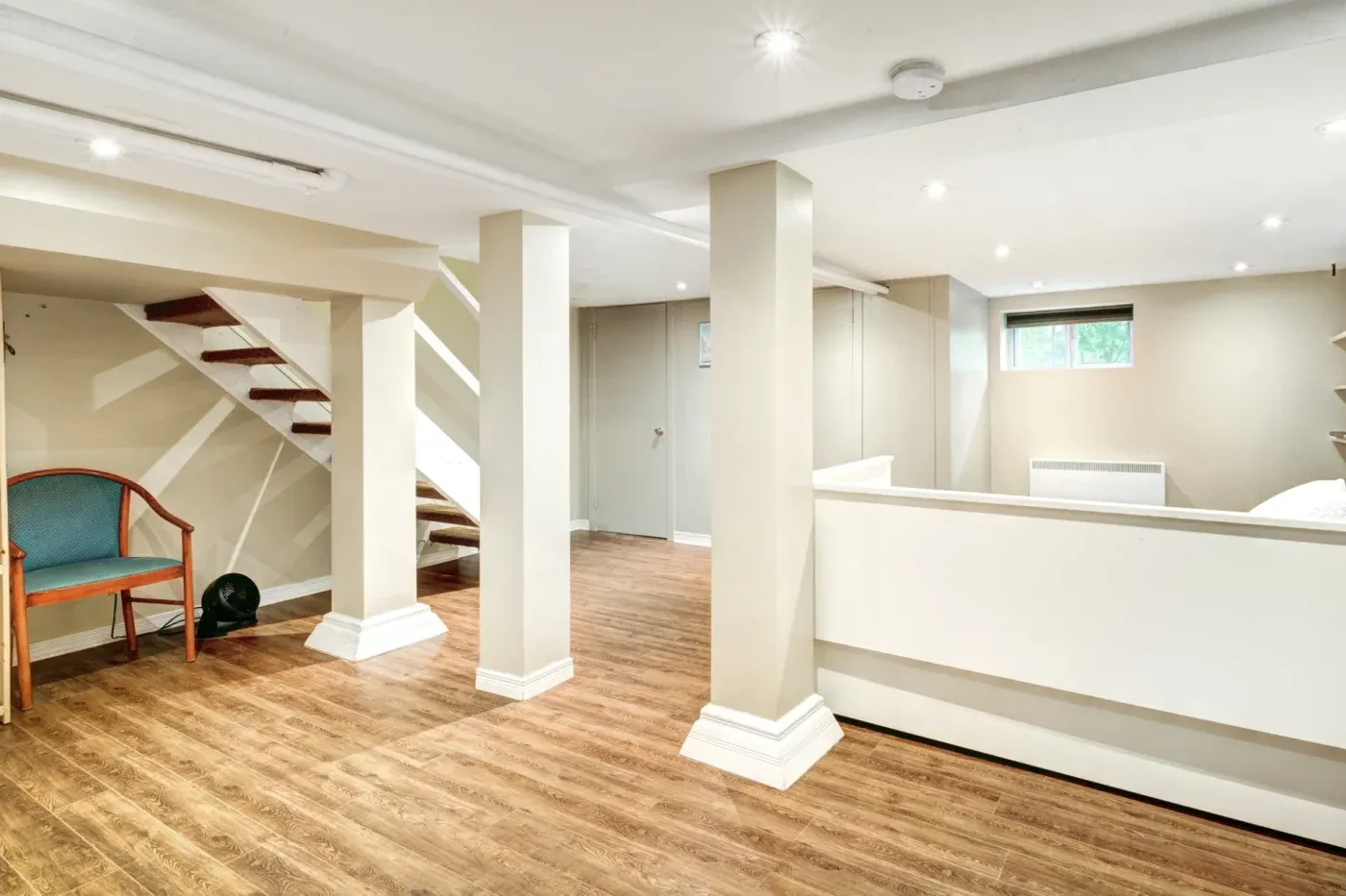 Spacious, well-lit basement with wooden flooring, white pillars, and a small window. A wooden chair is near a staircase, creating a calm, open atmosphere.