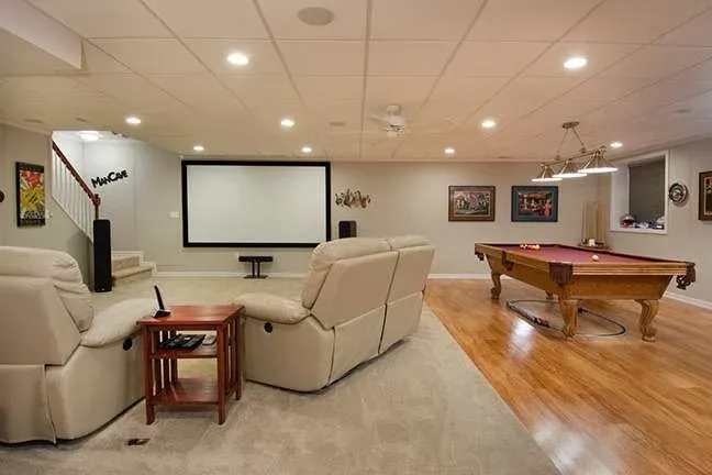 Basement game room features a pool table and large projector screen. Beige recliners face the screen. The room is warmly lit with cozy, relaxed ambiance.
