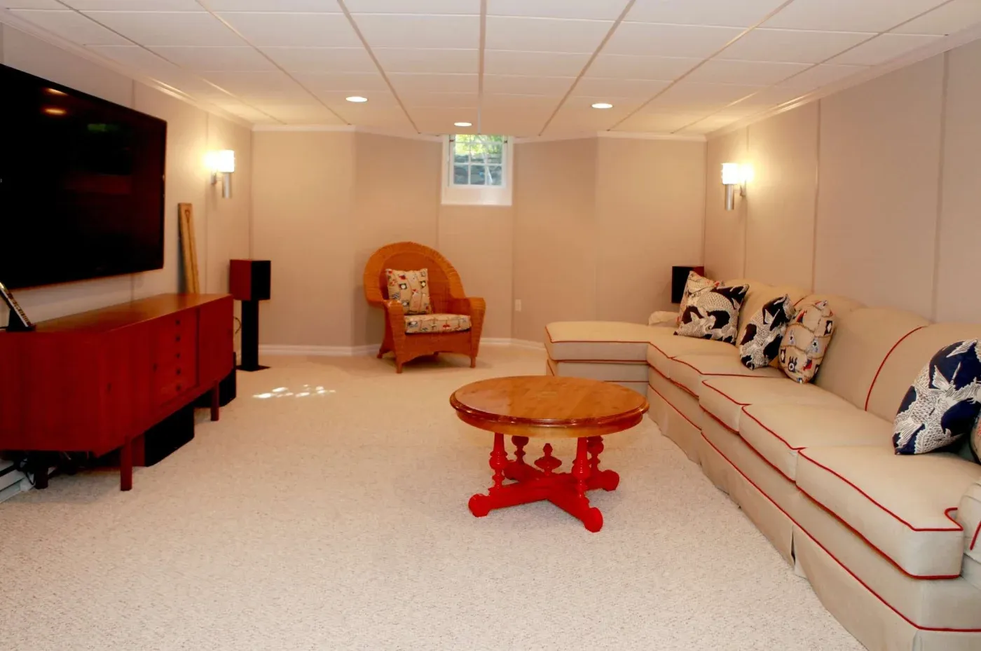 A cozy basement living room with beige walls, large sectional sofa adorned with patterned pillows, a red oval coffee table, a wicker chair, and a wall-mounted TV.