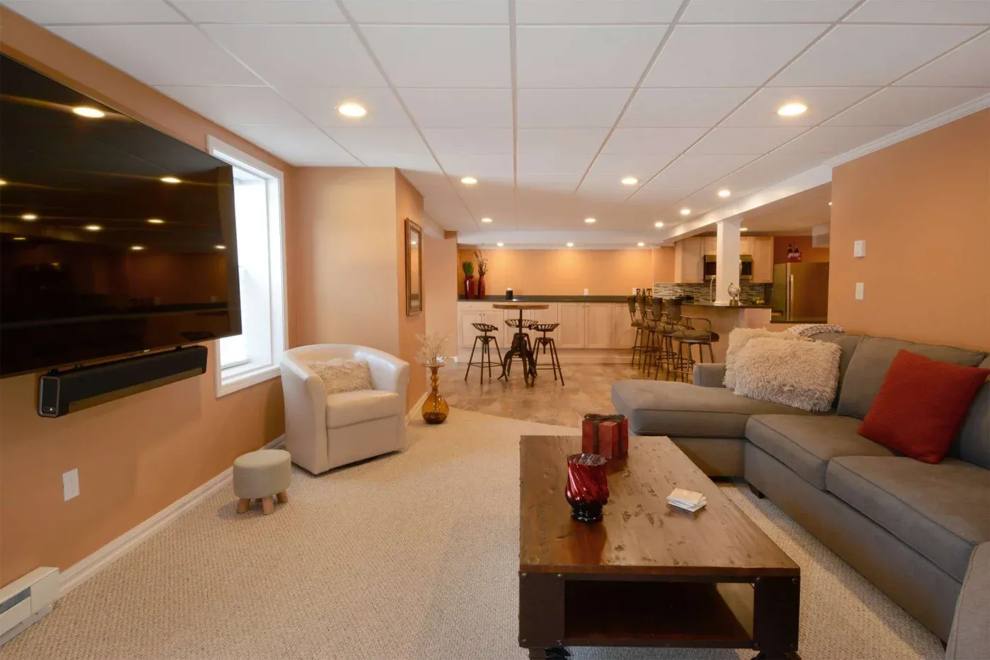 Spacious basement lounge with beige walls and ceiling lights. Features a large sectional sofa, wall-mounted TV, small bar with stools, and cozy atmosphere.