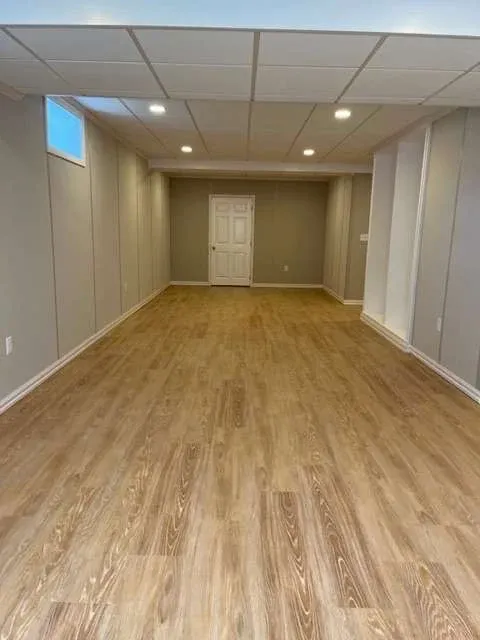 A long, narrow basement room with wood flooring, gray walls, and a white door at the far end. Recessed lighting creates a clean, inviting space.