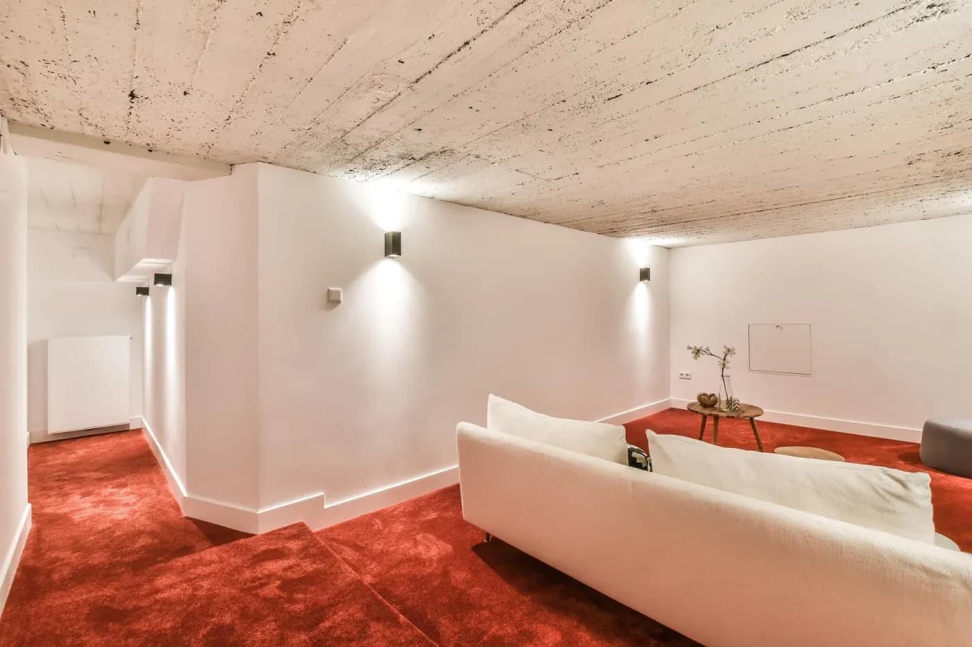 Minimalist basement room with low, textured ceiling and white walls. Soft lighting casts gentle shadows. Red carpet contrasts with a white sofa. Cozy and serene atmosphere.