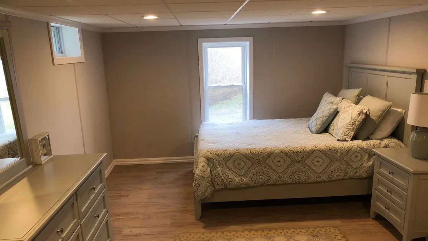 A cozy bedroom with beige walls and wooden flooring. A neatly made bed with patterned bedding is near a window, alongside a matching white dresser and nightstand.