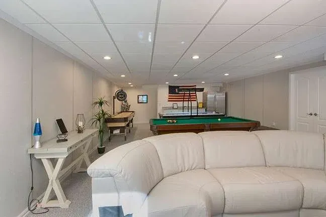 Spacious basement game room with beige walls and ceiling. Features a pool table, a beige sectional sofa, a foosball table, and an American flag on the wall. Cozy and inviting.