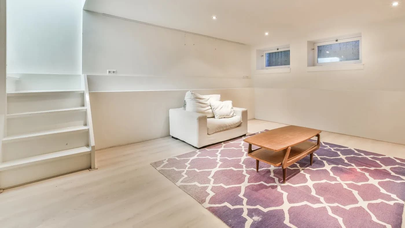 Minimalist basement room with white walls, a single white armchair, and a wooden coffee table on a purple geometric rug. Soft lighting and two small windows create a cozy ambiance.