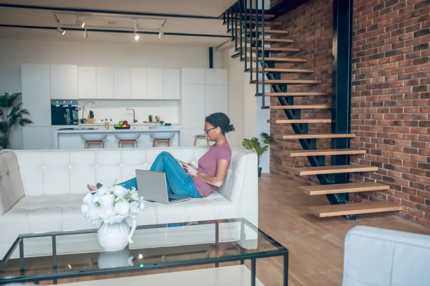 A woman sits on a white sofa with a laptop on the cushion, in a modern loft with a brick wall and spiral staircase. The room feels calm and cozy.