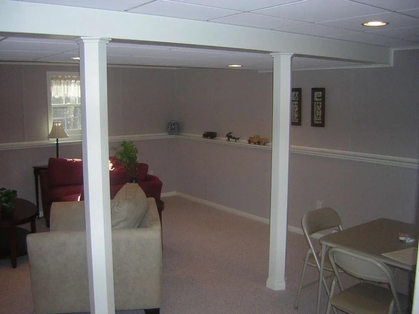 Spacious room with beige walls and carpet, featuring a red sofa, beige chair, folding table with chairs, and soft lighting from a nearby window. Cozy and inviting.