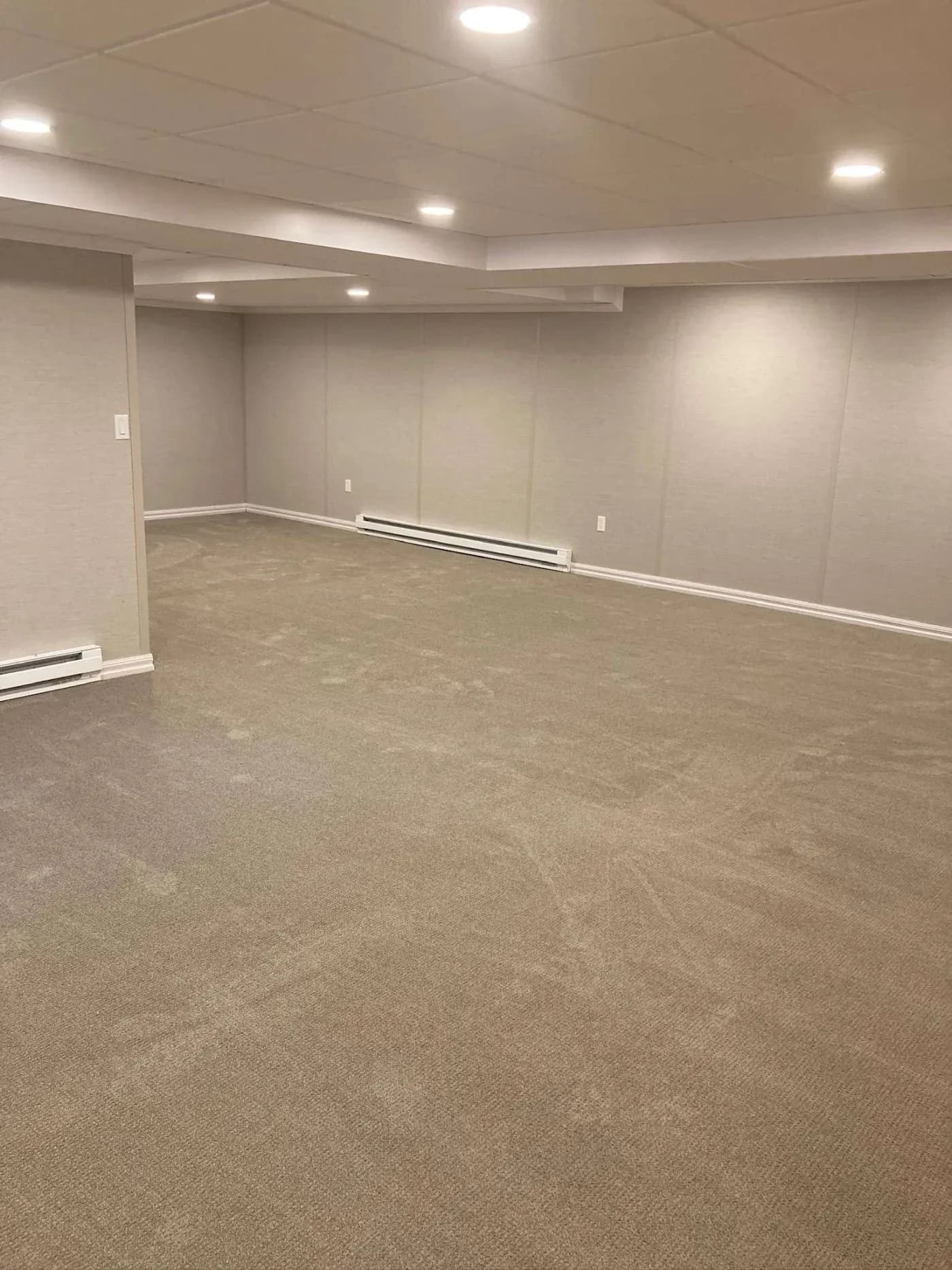 A spacious, empty basement room with gray carpeting and beige walls. The ceiling features recessed lighting, creating a bright and clean atmosphere.