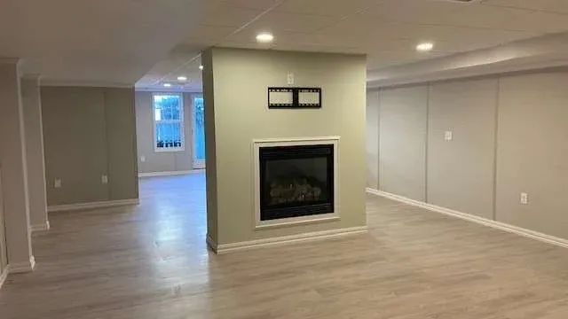 Spacious basement with light wood flooring, centered fireplace, neutral beige walls, recessed lighting, and a glass door leading outside. Cozy and modern.