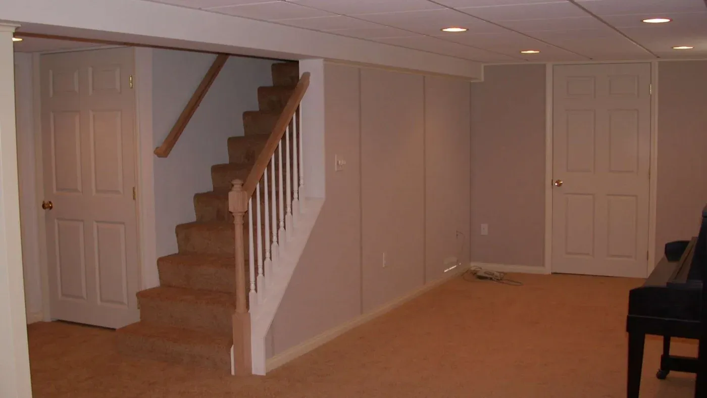A carpeted basement with neutral beige walls and recessed lighting. A staircase with wooden railings leads up, and a door is visible on the left. The space is clean and orderly.