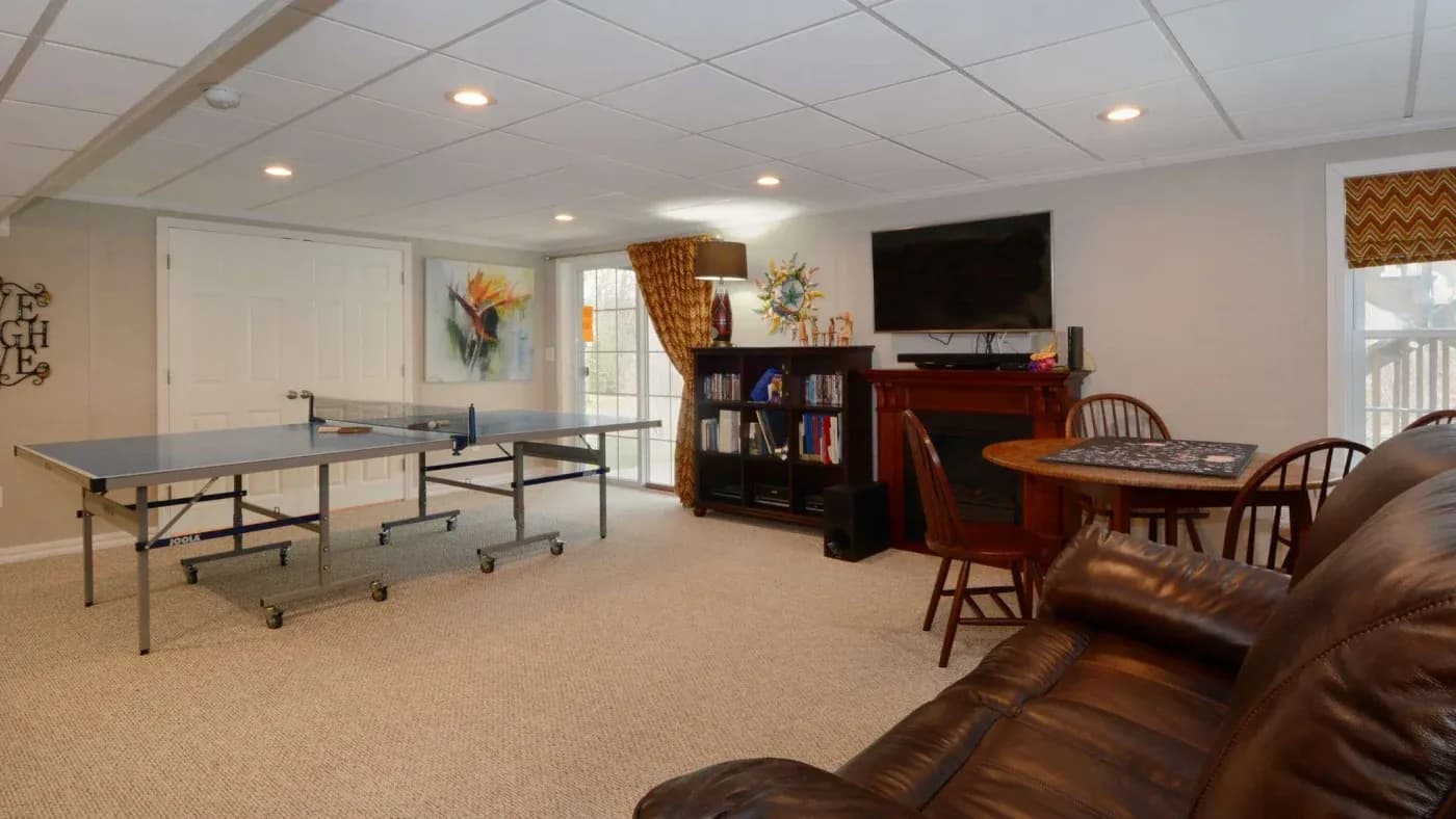 A cozy basement recreation room with a leather sofa, a ping pong table, a round wooden table with chairs, and a TV above a fireplace. Neutral walls and warm lighting create a welcoming atmosphere.