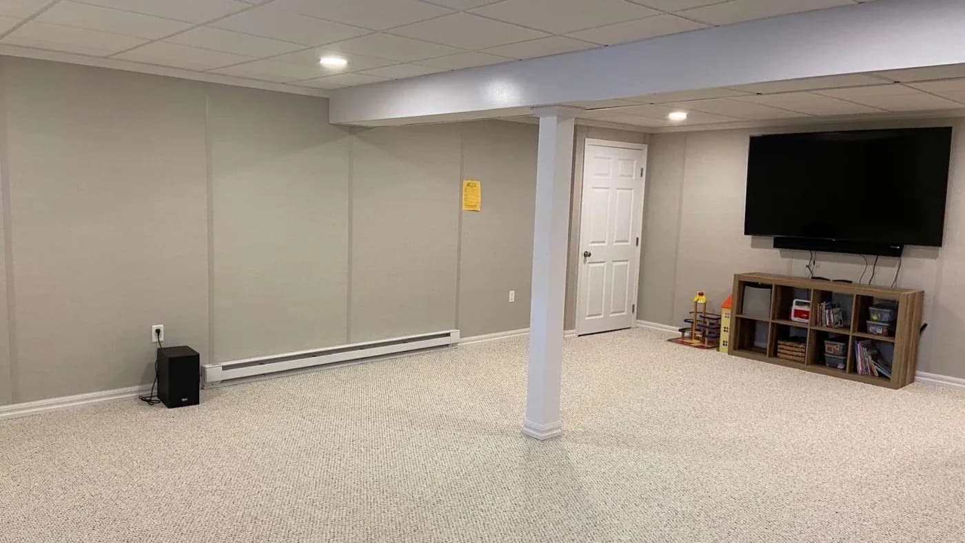 Spacious basement room with light gray walls and carpet. A large TV is mounted on the right wall above shelves with books and toys. A column stands centrally.