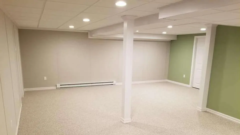 A spacious, empty basement with light beige walls and a green accent wall on the right. It has a white ceiling, recessed lighting, and neutral carpet flooring.
