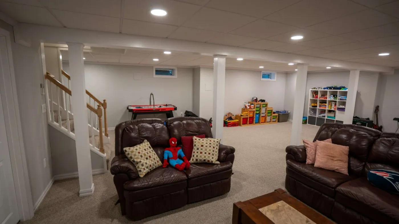 A spacious basement playroom with brown leather couches, a red and blue superhero toy, shelves with colorful bins, and an air hockey table, conveying a cozy and fun atmosphere.