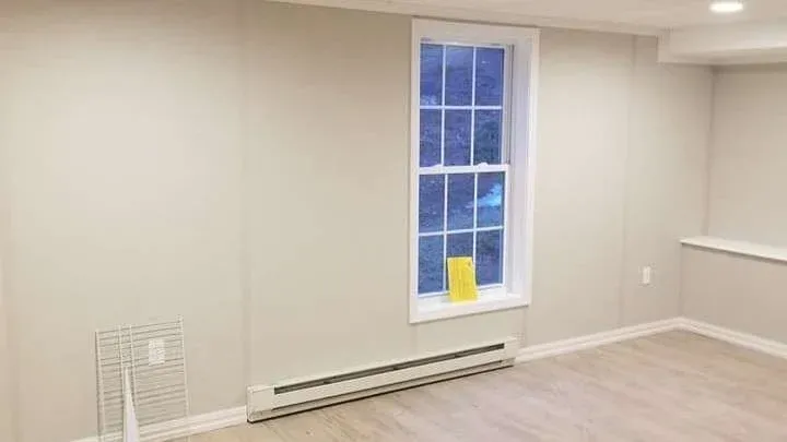 A minimalistic room with light-colored walls and wooden floors. A large window shows a view outside, and a small yellow object rests on the windowsill. A heating element runs along the base of the wall.