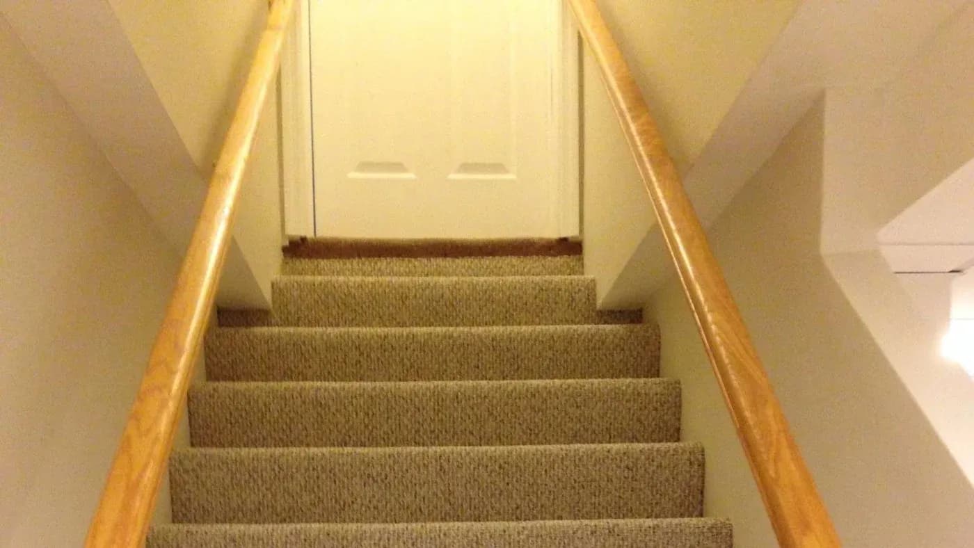 Carpeted stairs with wooden railings lead up to a white, closed door at the top. The lighting is warm, creating a cozy and inviting atmosphere.