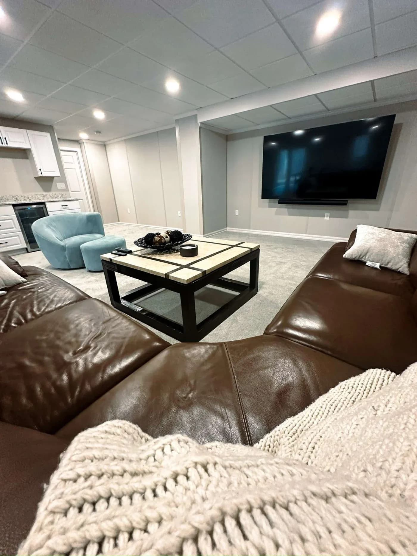 Cozy living room with large brown leather sectional, soft beige throw, sleek coffee table, blue accent chair, wall-mounted TV, and recessed lighting.