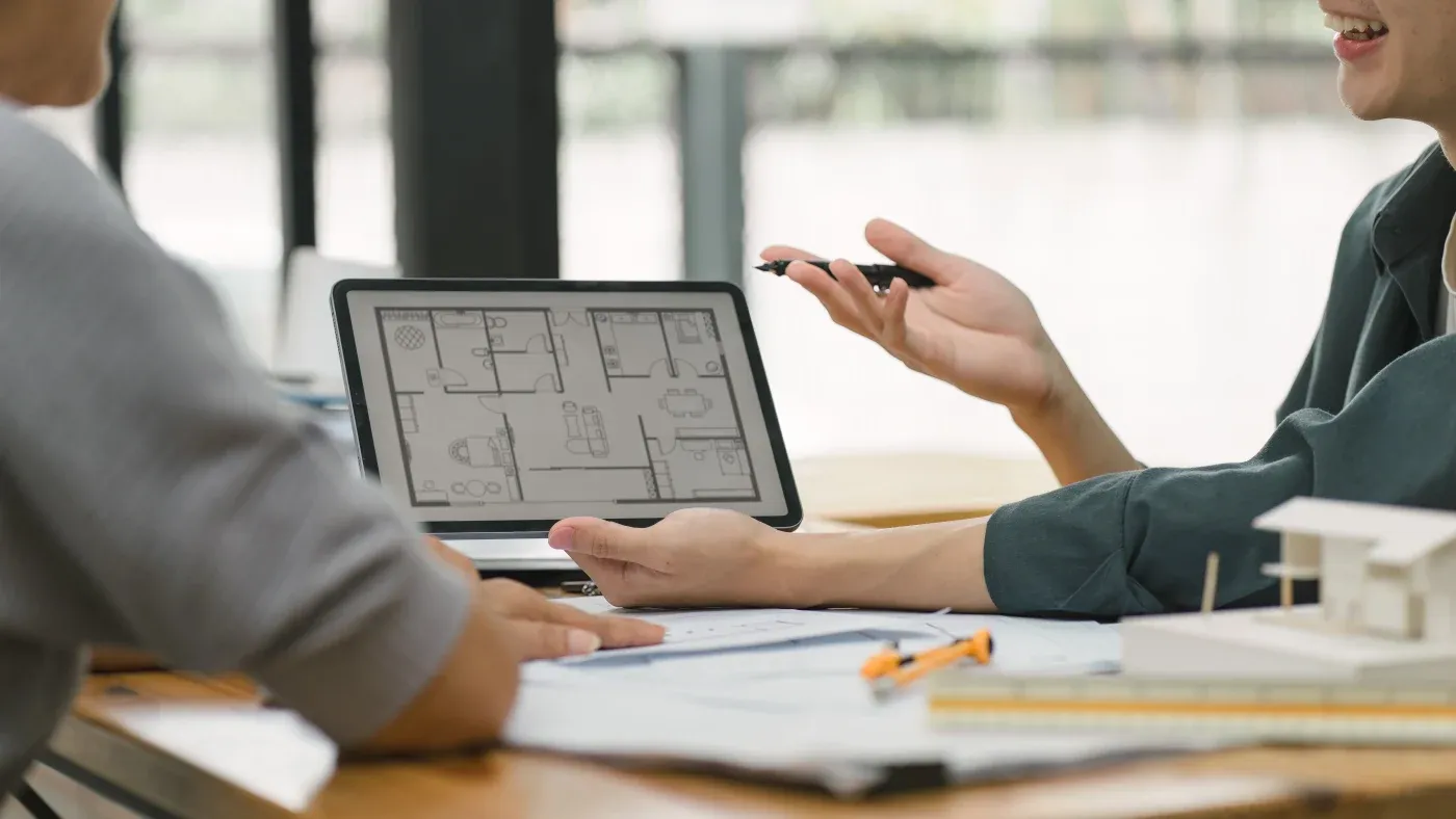 Two people discuss architectural plans on a tablet at a desk. One gestures with a pen, creating a collaborative and professional atmosphere.
