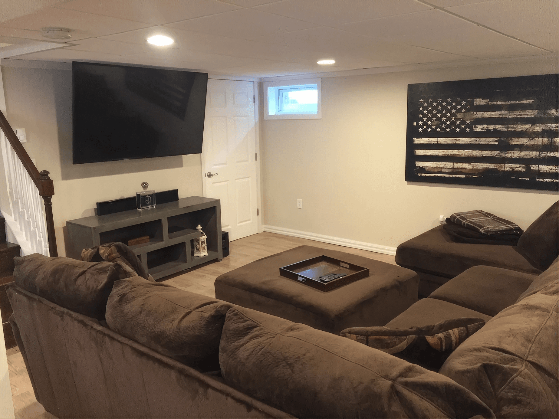 Finished basement family room featuring a large sectional sofa, oversized ottoman, wall-mounted TV, and modern entertainment console. Neutral finishes, wood-look flooring, recessed lighting, and American flag wall art create a cozy space for relaxing and watching movies.
