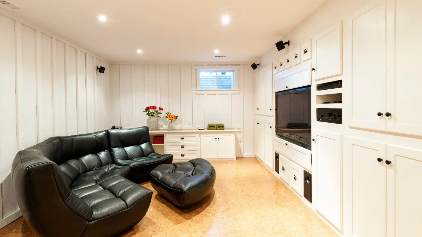 A cozy living room with white paneled walls, a large black leather sectional sofa, and a TV in a white built-in unit. Soft lighting creates a warm ambiance.