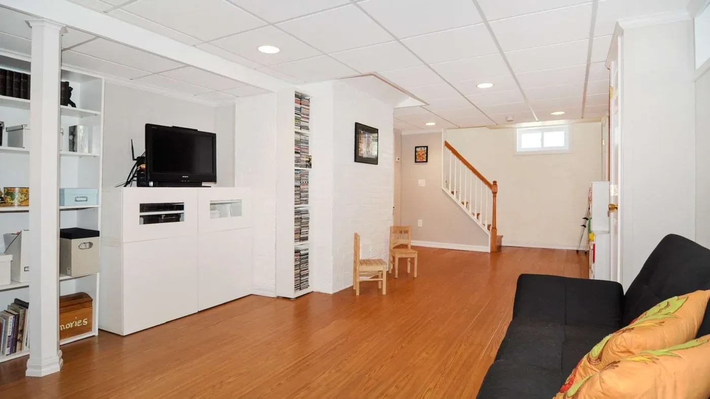 Spacious basement living area with wooden flooring, featuring a black sofa with orange pillows, TV on a white media unit, and a staircase in the background.