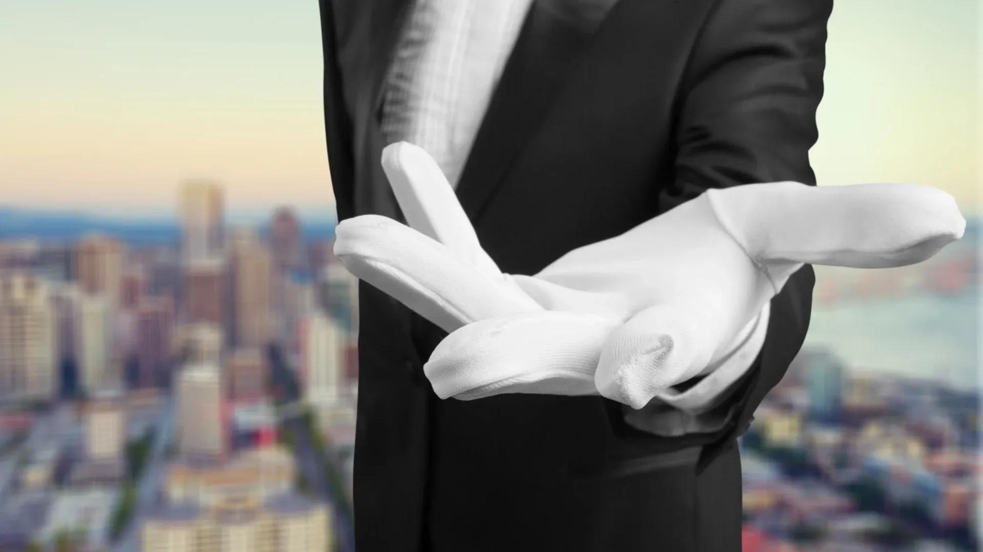 Person wearing a black suit and white gloves extends their hand invitingly towards the viewer, with a blurred cityscape in the background, suggesting elegance.