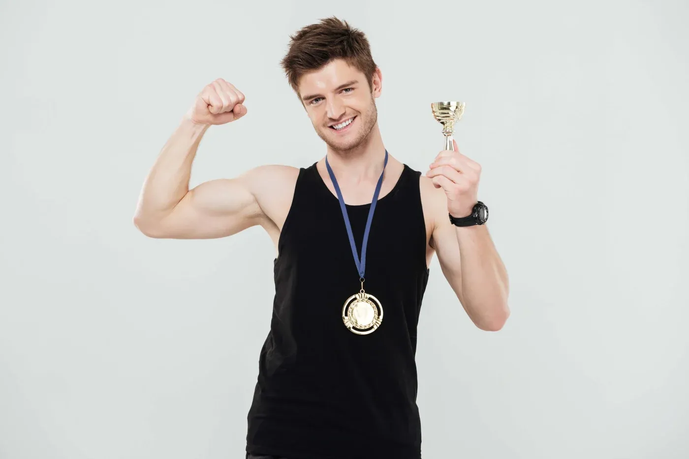 A man in a black tank top holds a trophy and wears a gold medal, smiling confidently. He flexes his arm, showing a sense of achievement and pride.