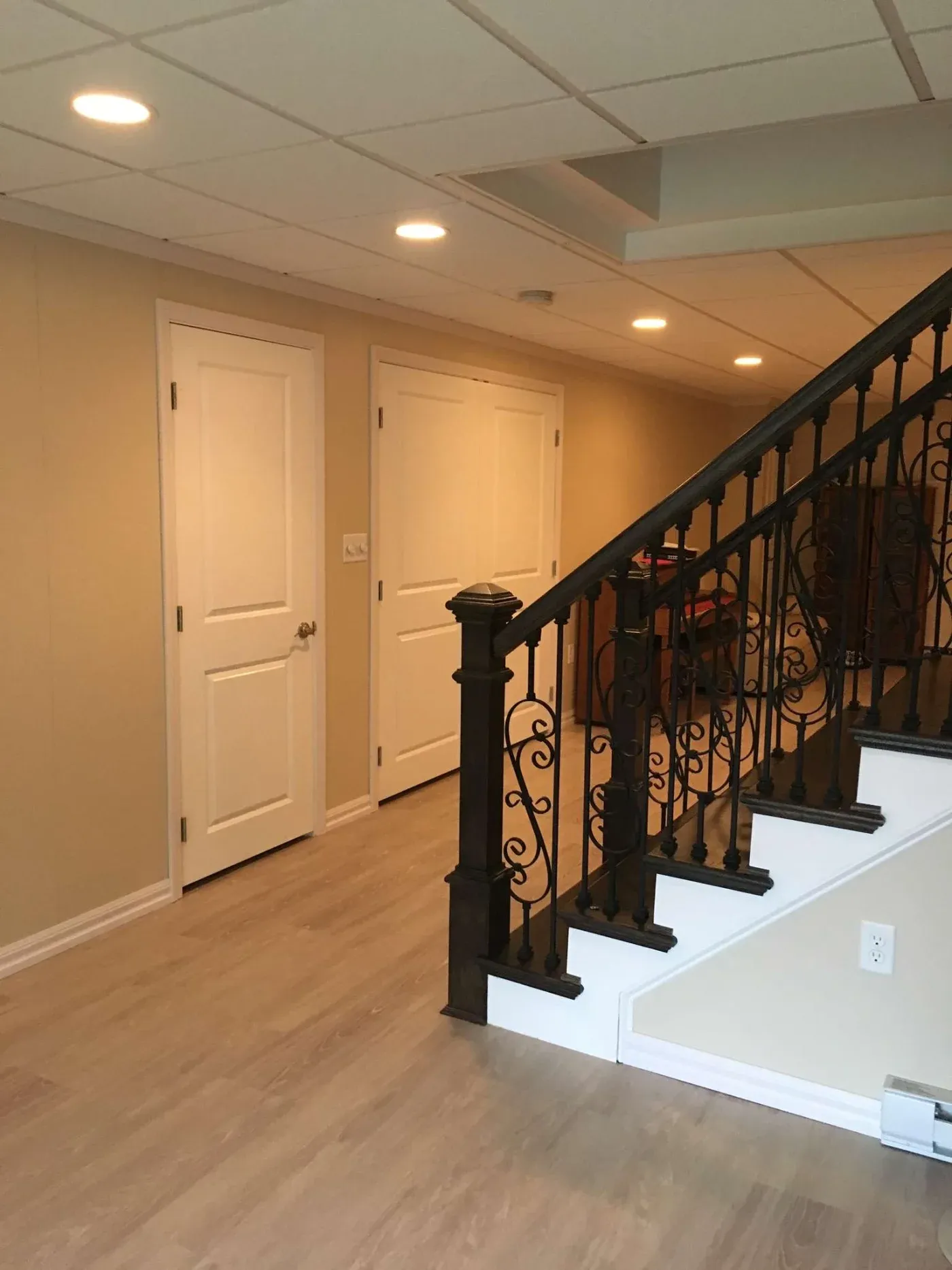 Basement with beige walls, light wood flooring, and white doors. A dark ornate wrought iron staircase leads to upper floor, creating a cozy ambiance.