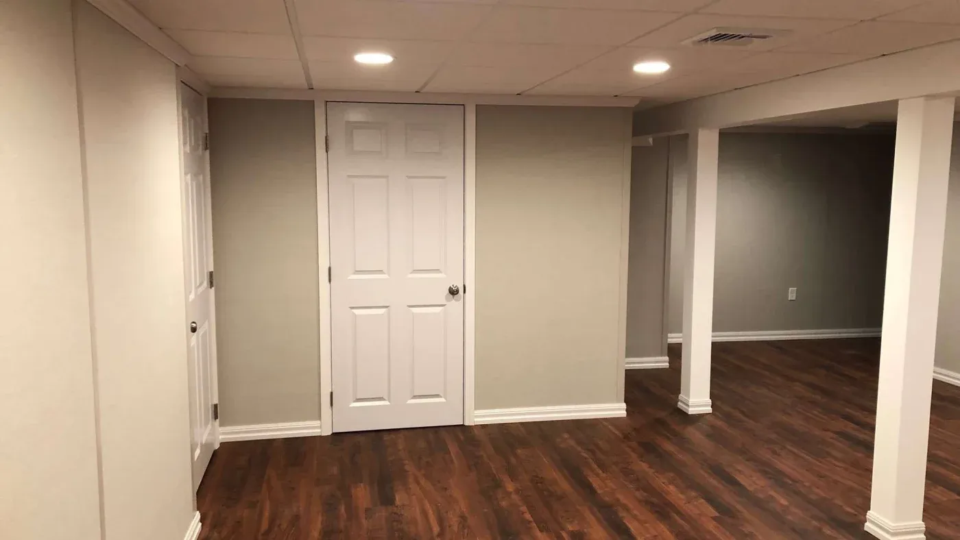 A finished basement with hardwood floors, beige walls, and a white door. Recessed lighting illuminates the space, creating a clean, modern feel.
