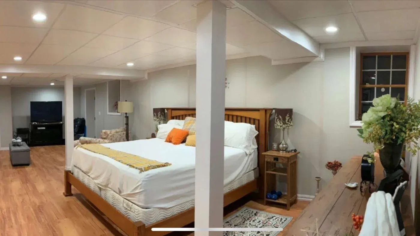 A cozy basement bedroom with a wooden bed, white and orange bedding, and decorative pillows. Warm lighting, hardwood floors, and a flat-screen TV create a welcoming ambiance.