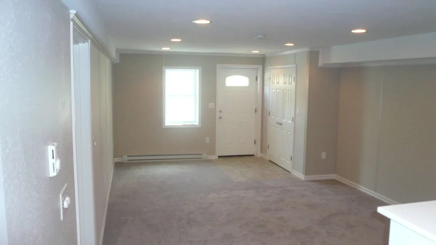 Spacious room with beige walls and carpet, featuring a white entrance door and window. Recessed ceiling lights illuminate the simple, welcoming space.