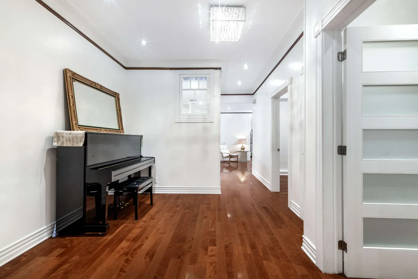 A bright hallway with polished wooden floors features a black piano with a bench beneath a large ornate mirror. A modern chandelier hangs above.