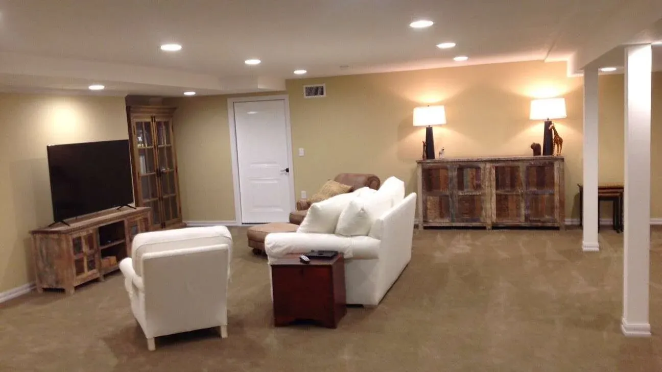 A cozy basement living room with beige walls and carpet. It features white sofas, a large flat-screen TV, a wooden cabinet, and soft lighting from table lamps, creating a warm atmosphere.