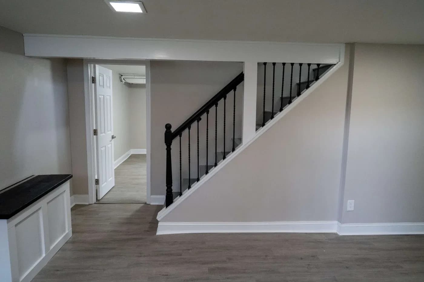 Basement room with light gray walls and wood flooring. A black handrail staircase is on the right, and a door leads to another room on the left. Modern and clean.