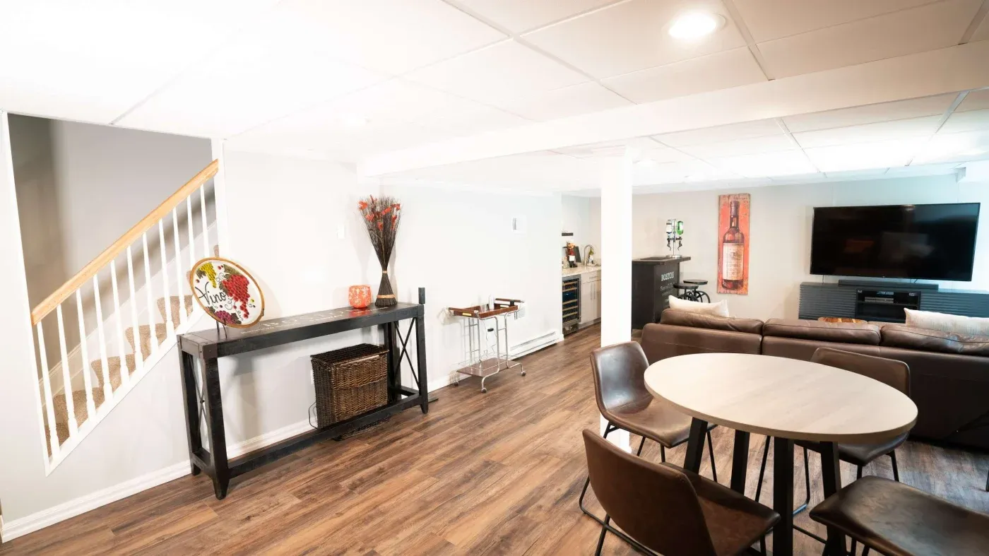 Modern basement with wood flooring, featuring a round dining table, brown chairs, a gray sectional sofa, wall art, and a large flat-screen TV.