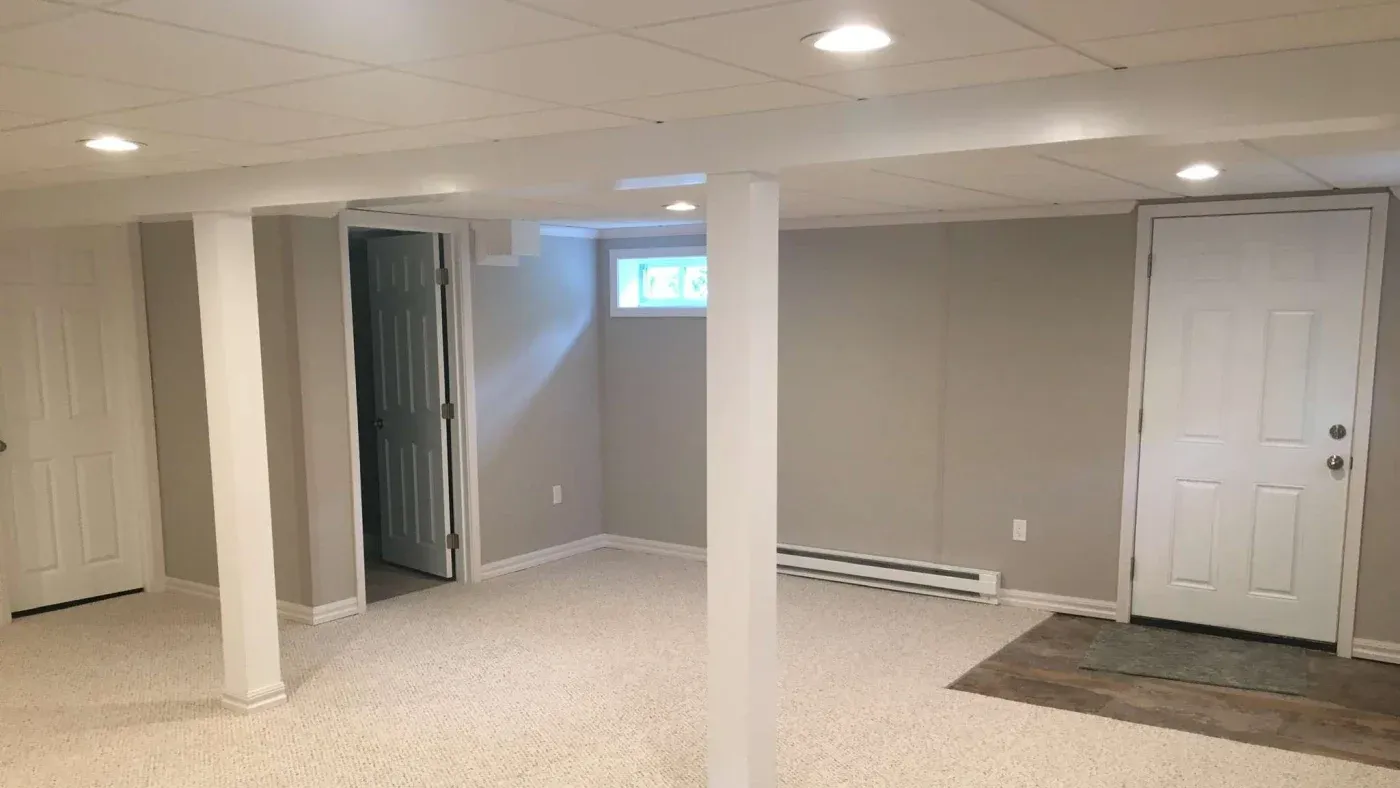 A spacious basement with neutral walls, carpeted floor, ceiling lights, and support columns. There are two doors and a small window, creating a clean and inviting atmosphere.