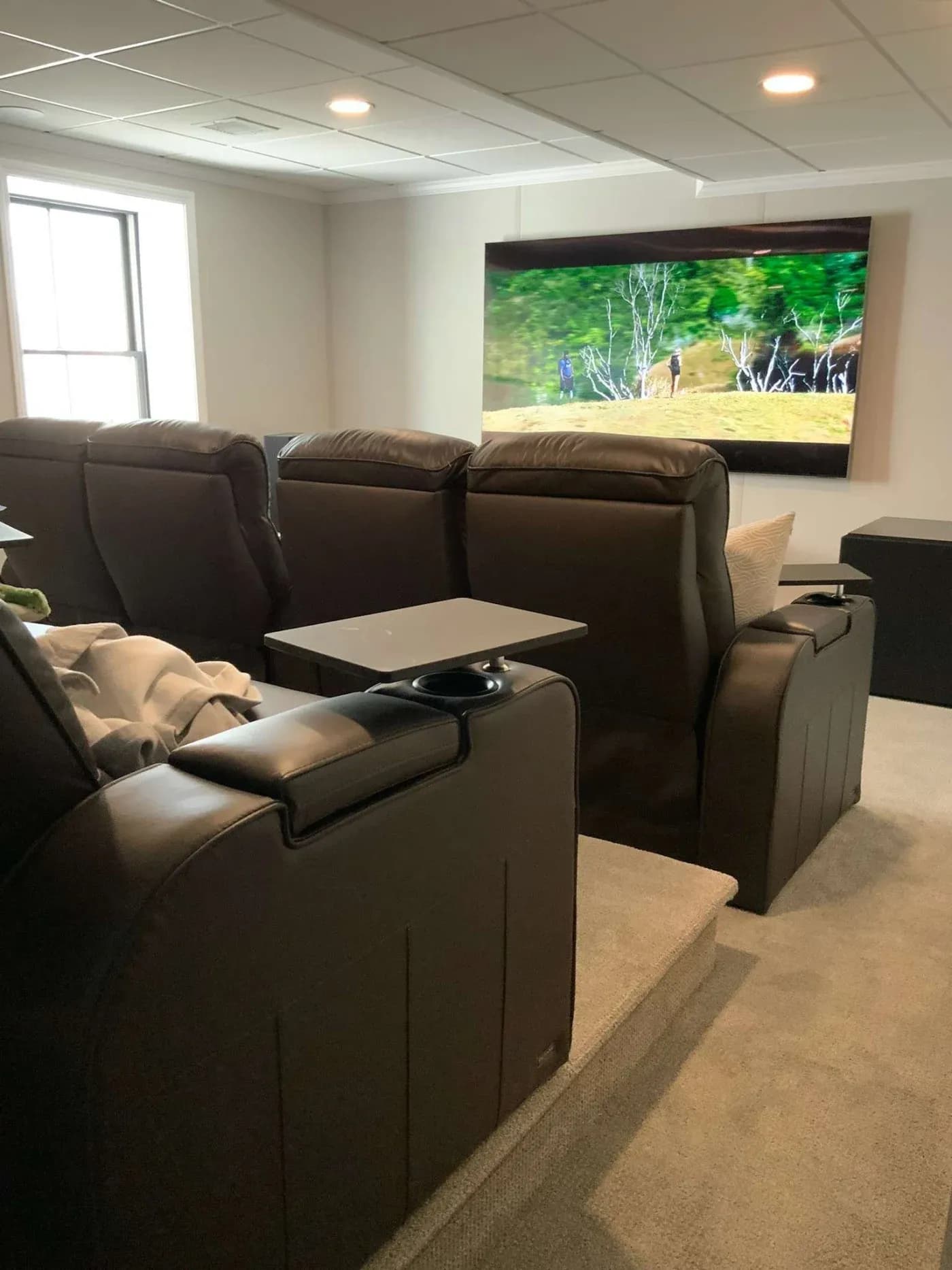 A cozy home theater with brown leather recliners facing a large wall-mounted screen showing a nature scene. Soft lighting creates a relaxing ambiance.