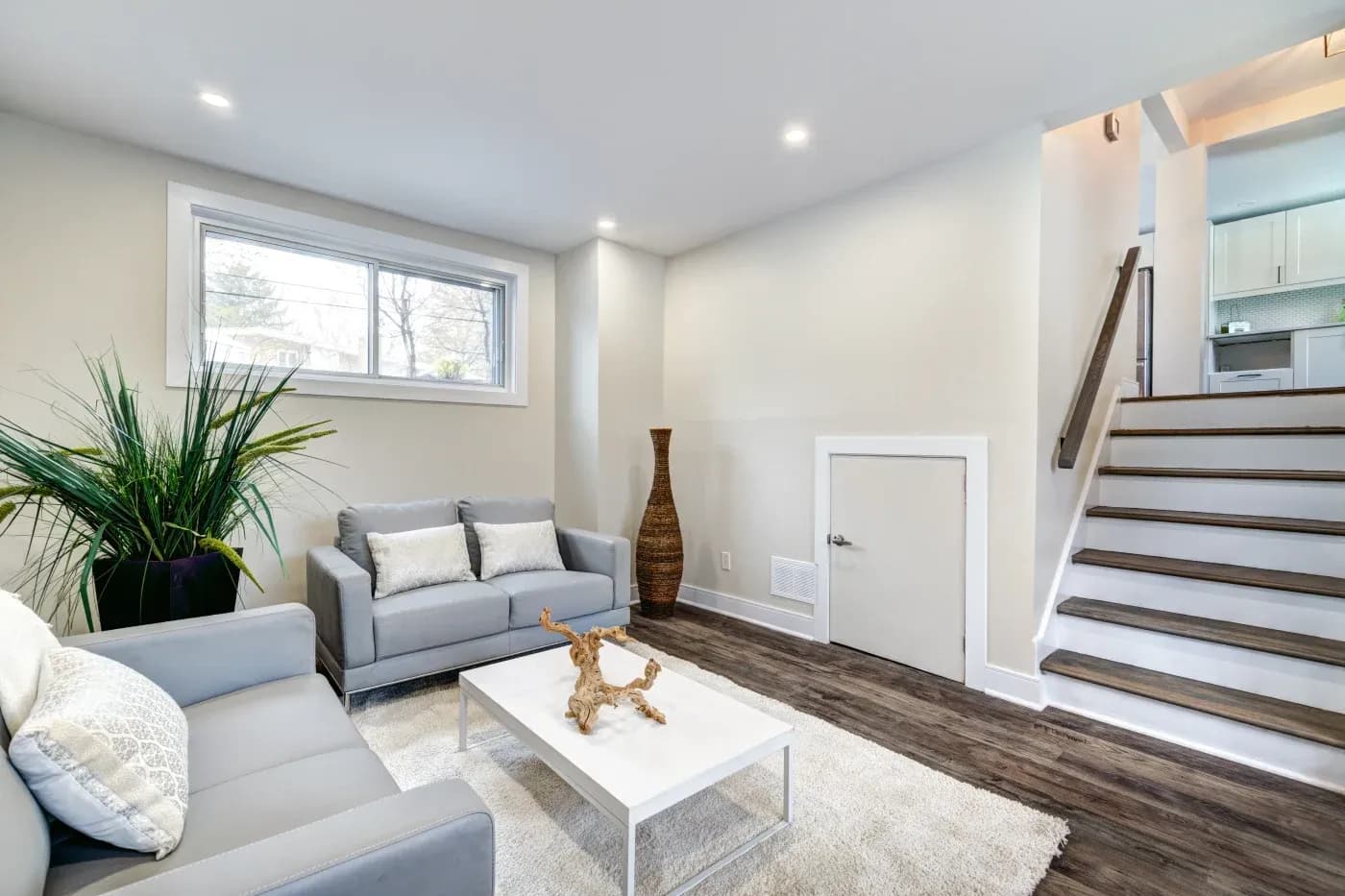 Modern living room with gray sofas, white cushions, a white coffee table, and a decorative wood centerpiece. A large plant is by the window, and a tall vase stands near the staircase. The space is bright and inviting.