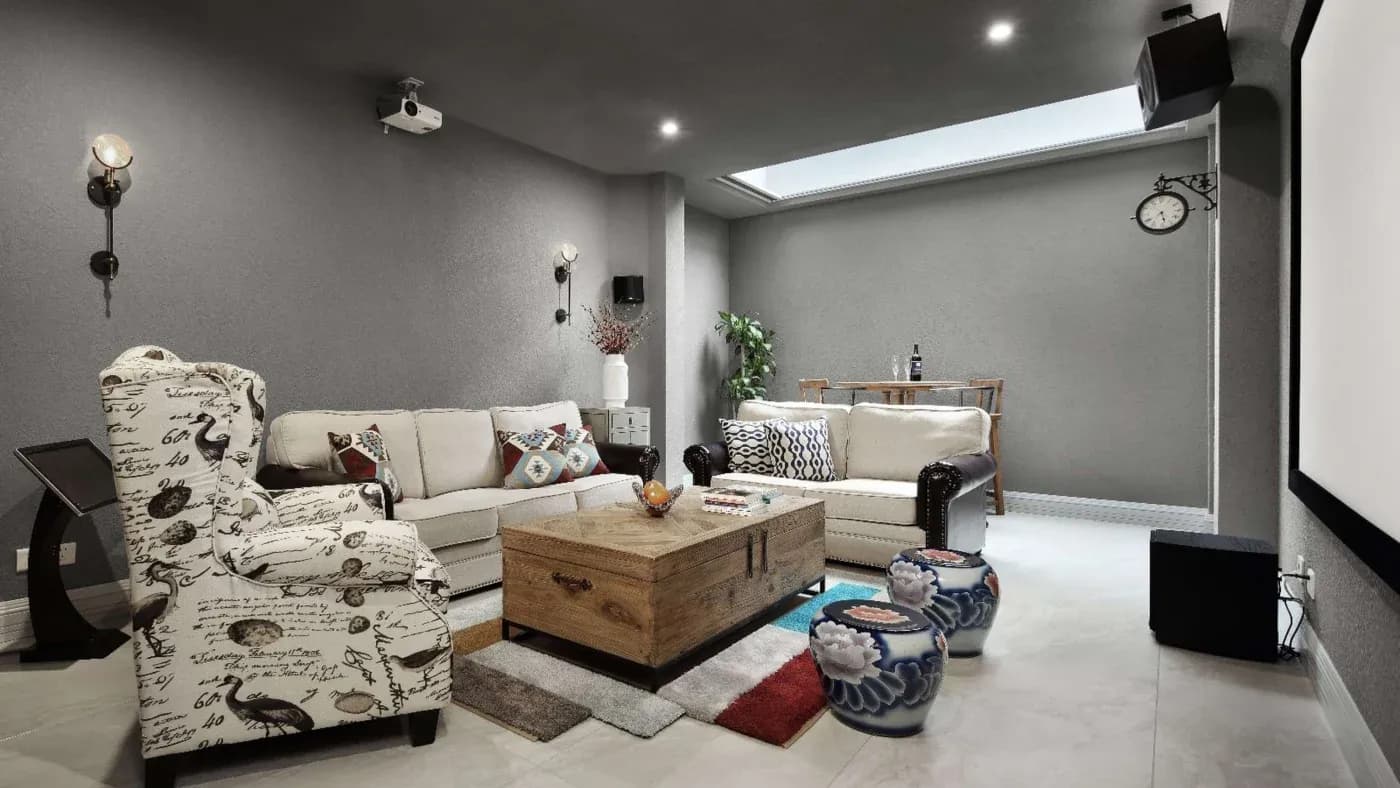 A cozy, modern living room with gray walls, two white sofas, a patterned armchair, a wooden coffee table, and colorful cushions. There's a large screen, projector, and decorative items like a large clock and plants, creating a warm and inviting ambiance.