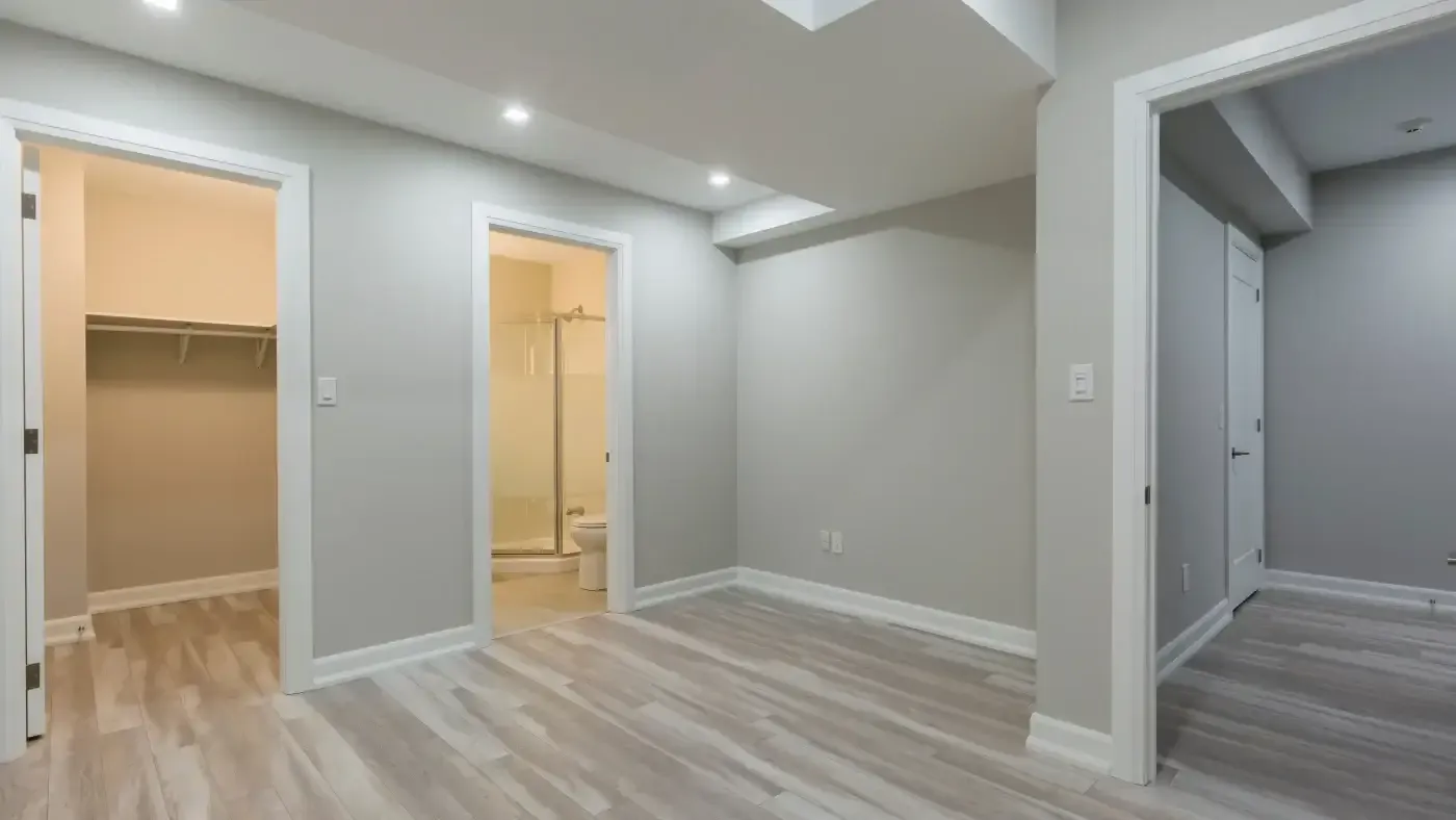 A modern, empty room with light gray walls and wooden flooring. It features a walk-in closet and a bathroom with a glass shower. Recessed lighting adds brightness.
