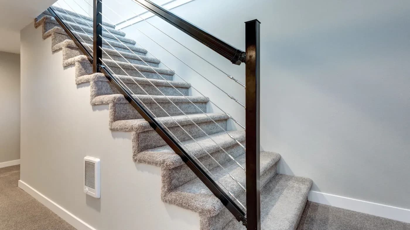 A modern staircase with gray carpeted steps and sleek steel cable railing against a light gray wall, creating a clean and contemporary feel.