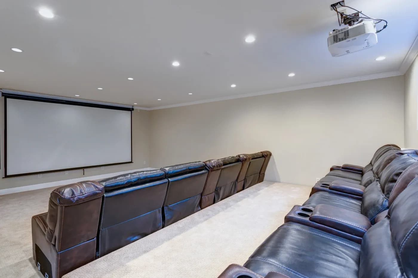 A spacious home theater with two rows of plush brown leather recliners facing a large projection screen. The room is well-lit with ceiling lights.