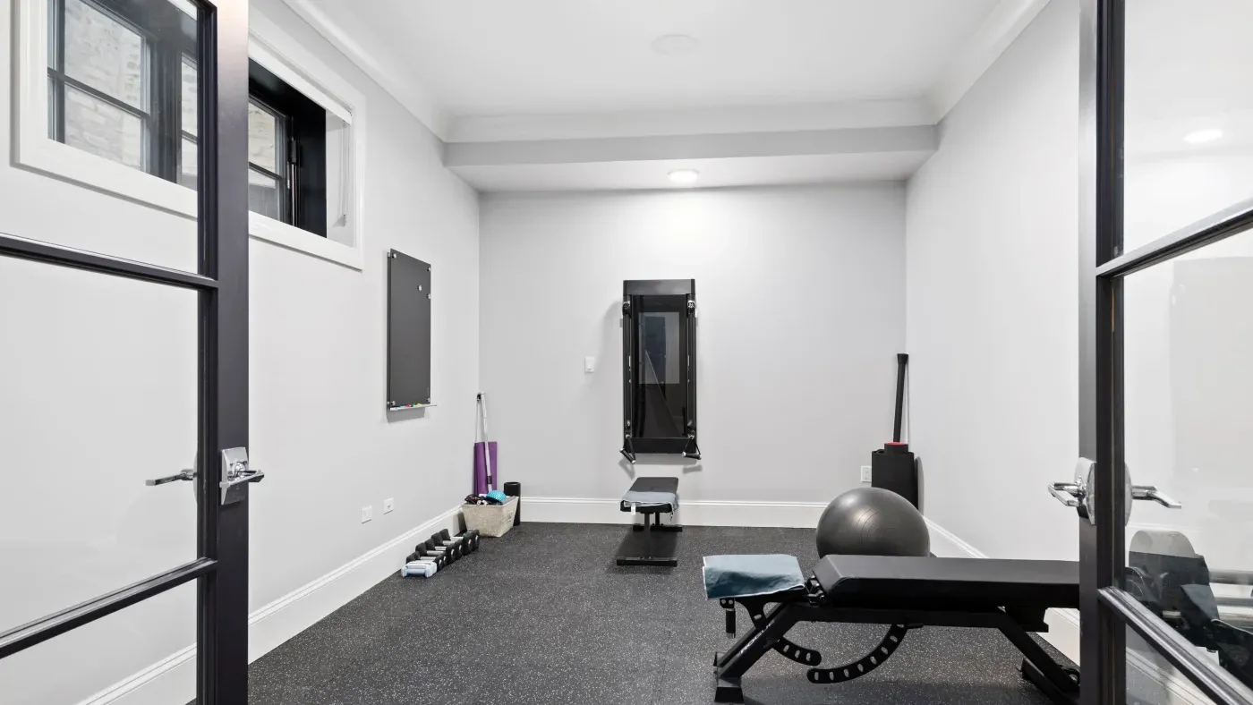A minimalist home gym with white walls and black flooring features a weight bench, exercise ball, resistance bands, and a wall-mounted workout machine. The space is well-lit and inviting.
