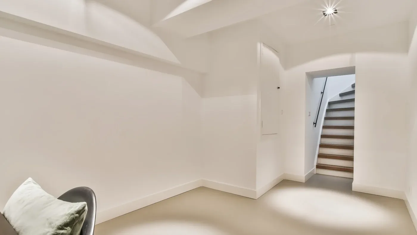 A minimalist, well-lit room features a neutral-tone chair with a white cushion. A staircase with wooden steps leads up, creating an airy, calm atmosphere.