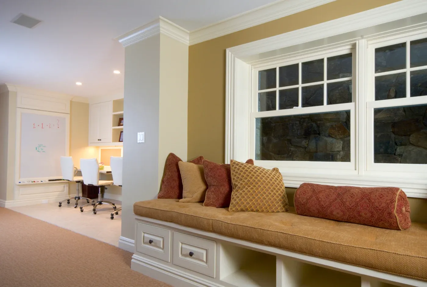 Cozy room with a window seat adorned with patterned cushions, beige walls, and a view of a home office featuring a whiteboard and chairs.