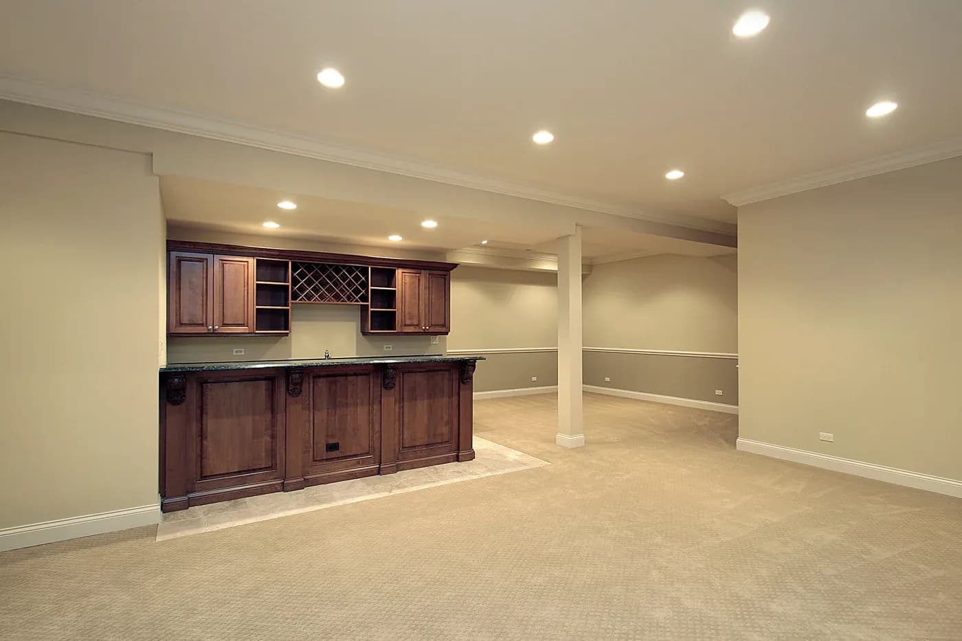 Spacious basement with beige walls and carpet, featuring a mahogany bar with wine storage and overhead lighting, creates a warm, inviting ambiance.